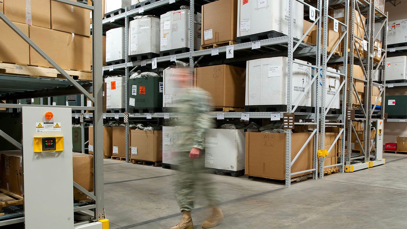 soldier checking gear on a table in a warehouse with high bay storage system behind him