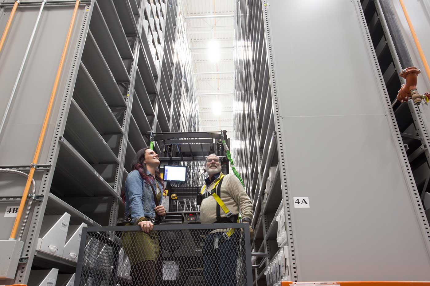 Two people on a lift in between two high-bay shelving stacks